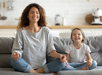 Mutter und Tochter praktizieren gemeinsam Achtsamkeit und Meditation zu Hause – beide sitzen entspannt im Schneidersitz auf dem Sofa mit geschlossenen Augen