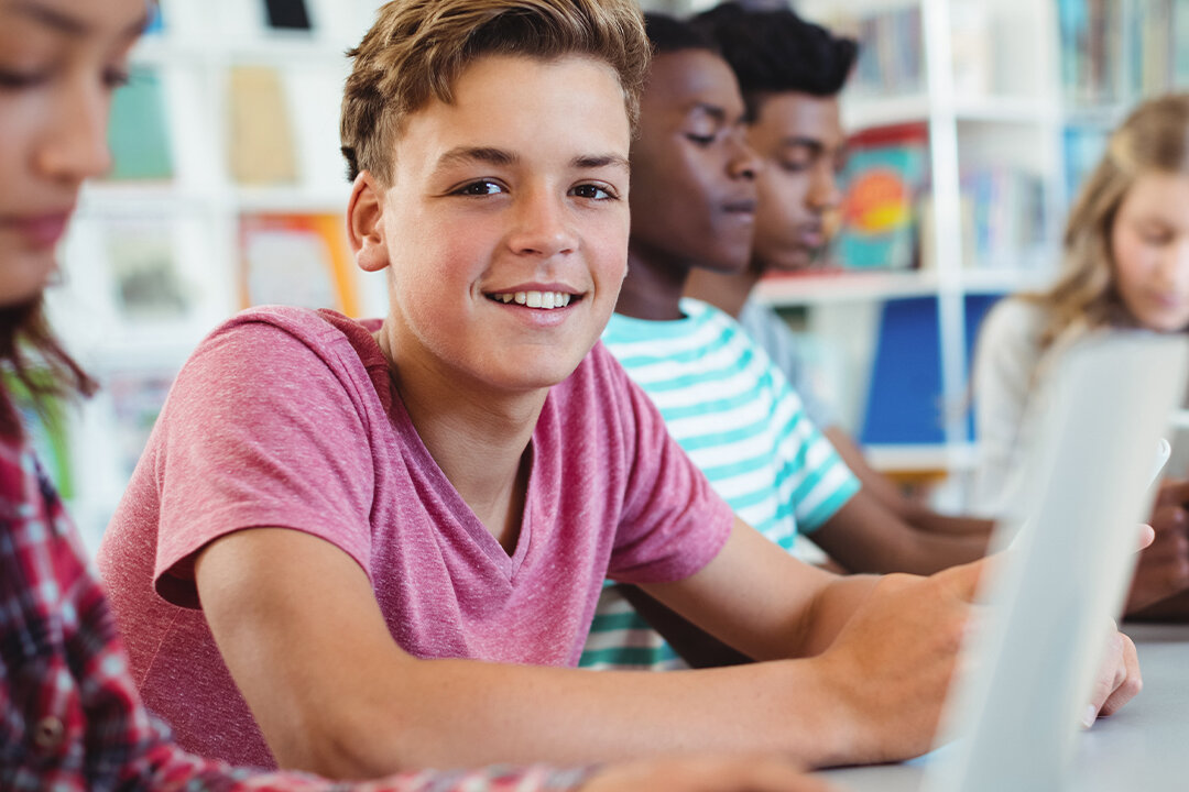 Ein Junge mit braunen Haaren und rosa T-Shirt lächelt in die Kamera. Im Hintergrund arbeiten weitere Kinder unterschiedlicher Herkunft gemeinsam an einem Tisch in einem hellen, modern eingerichteten Klassenzimmer. Das Bild illustriert positives, gemeinsames Lernen im Schulalltag.