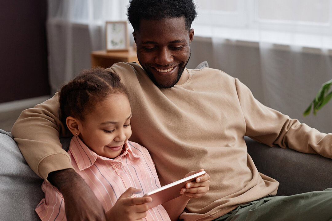 Vater und Tochter schauen lächelnd gemeinsam auf ein Tablet im Wohnzimmer.