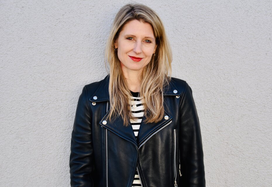 Laura Fröhlich, Redakteurin, Porträt mit warmem Lächeln und direktem Blick – schulterlange braune Haare mit Highlights, roter Lippenstift, schwarze Lederjacke über Streifenshirt, professionell und nahbar, Vertrauen und Expertise