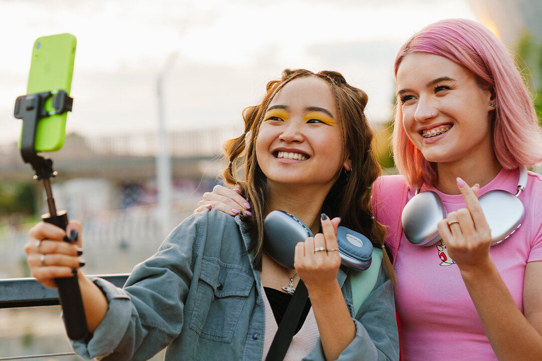 Zwei junge Frauen stehen nebeneinander, zeigen mit ihren Fingern ein Herz und posieren mit einem Smartphone auf einem Selfie-Stick.