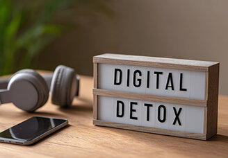 Leuchtbox mit Aufschrift „Digital Detox" auf Holzoberfläche, daneben abgelegtes Smartphone und Kopfhörer – Symbol für digitale Auszeit im Familienalltag