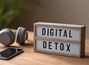 Leuchtbox mit Aufschrift „Digital Detox" auf Holzoberfläche, daneben abgelegtes Smartphone und Kopfhörer – Symbol für digitale Auszeit im Familienalltag