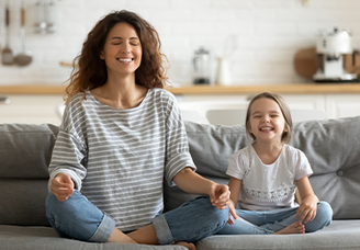 Mutter und Tochter praktizieren gemeinsam Achtsamkeit und Meditation zu Hause – beide sitzen entspannt im Schneidersitz auf dem Sofa mit geschlossenen Augen