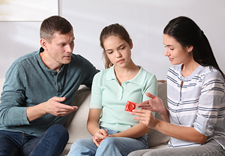 Zwei Erwachsene sprechen mit einem Mädchen auf dem Sofa und erklären ihr anhand eines Kondoms das Thema Verhütung
