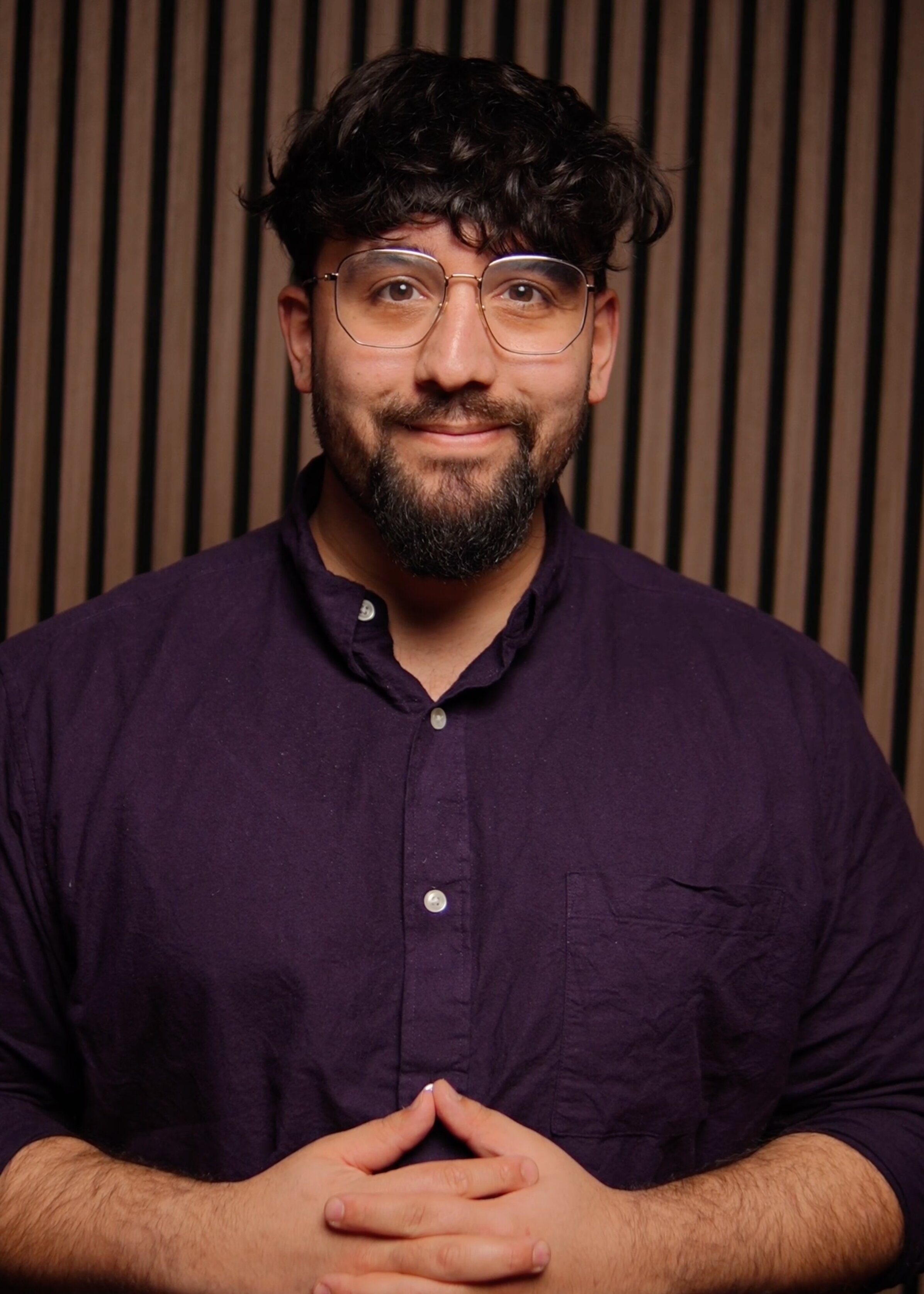 Lukas Maher, Redakteur, Porträt mit aufrichtigem Lächeln – dunkle, voluminöse Haare mit Textur, gepflegter dunkler Bart, klare Brille mit modernem Design, dunkles Navy-Hemd mit weißen Perlmuttknöpfen, Hände klassisch gefaltet, intellektuell und zugänglich, Vertrauen und Kompetenz