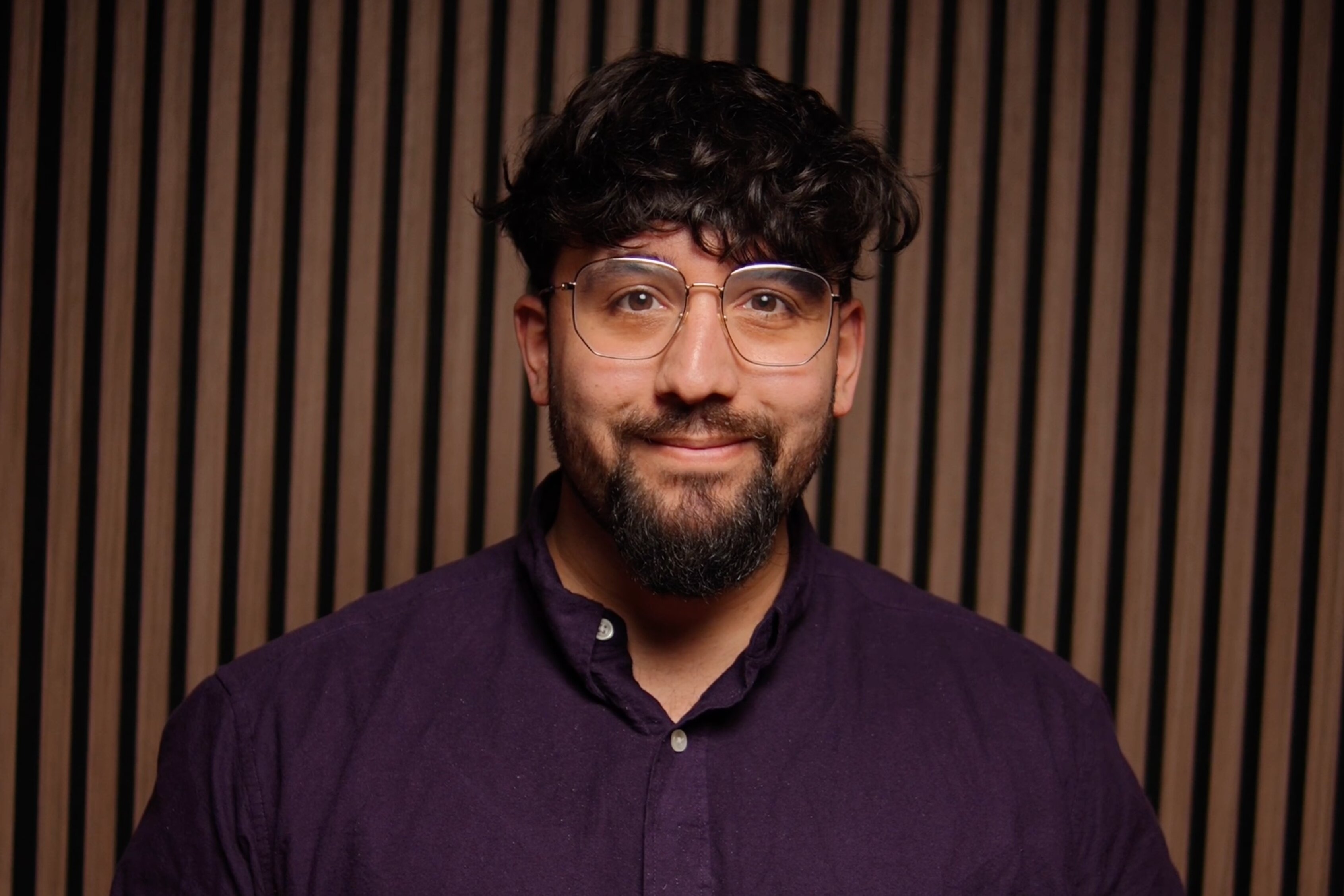 Lukas Maher, Redakteur, Porträt mit aufrichtigem Lächeln – dunkle, voluminöse Haare mit Textur, gepflegter dunkler Bart, klare Brille mit modernem Design, dunkles Navy-Hemd mit weißen Perlmuttknöpfen, Hände klassisch gefaltet, intellektuell und zugänglich, Vertrauen und Kompetenz