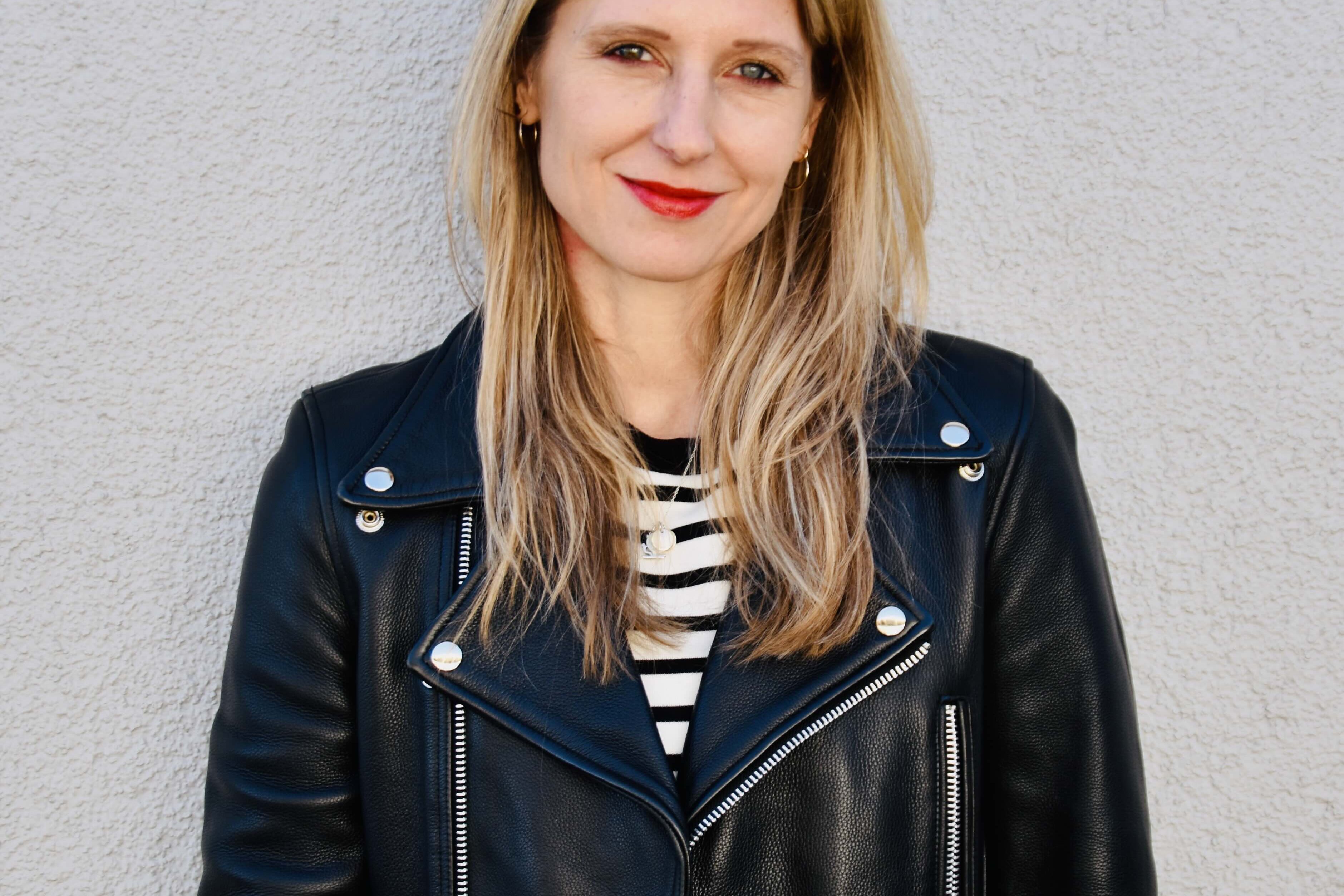 Laura Fröhlich, Redakteurin, Porträt mit warmem Lächeln und direktem Blick – schulterlange braune Haare mit Highlights, roter Lippenstift, schwarze Lederjacke über Streifenshirt, professionell und nahbar, Vertrauen und Expertise