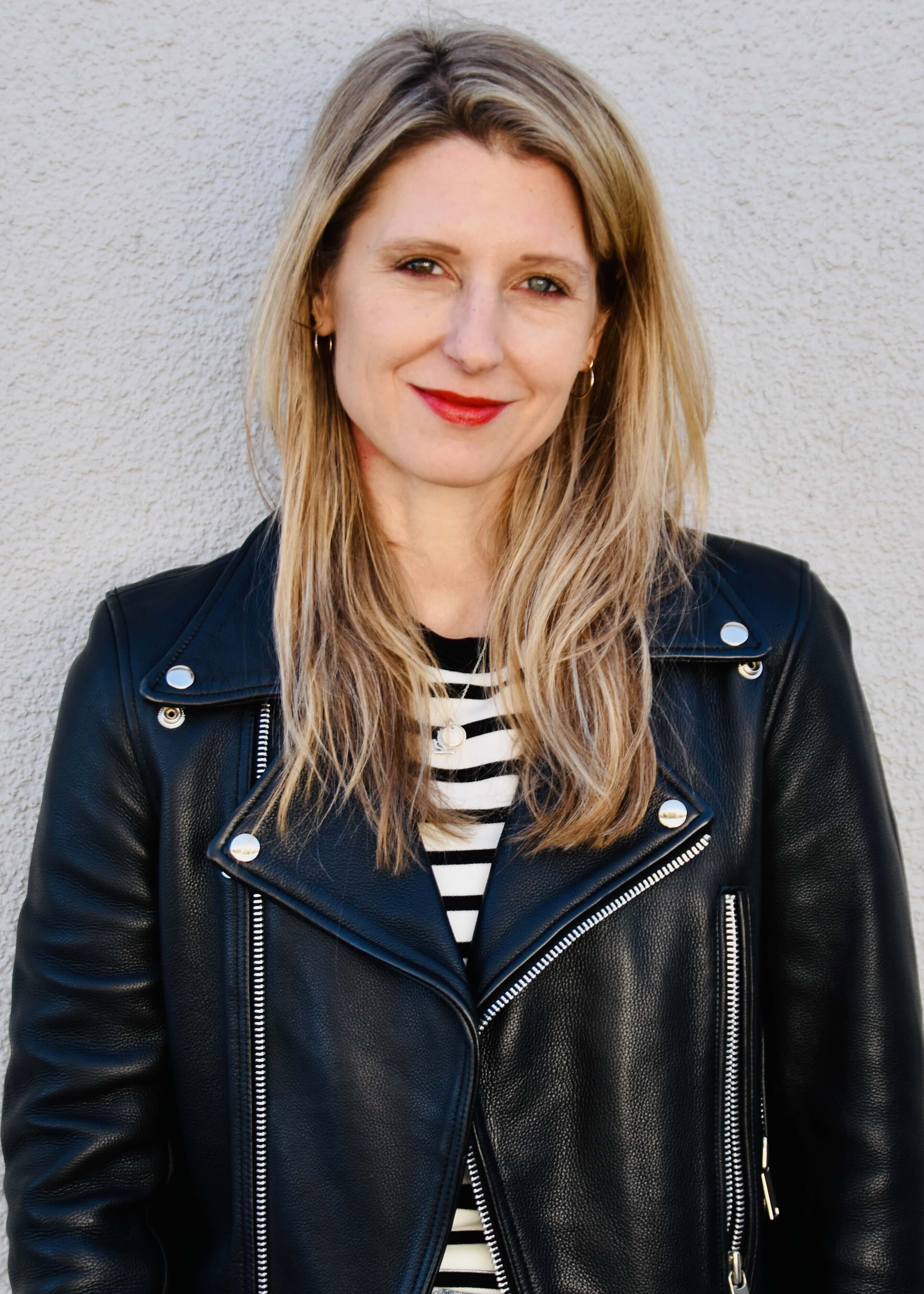 Laura Fröhlich, Redakteurin, Porträt mit warmem Lächeln und direktem Blick – schulterlange braune Haare mit Highlights, roter Lippenstift, schwarze Lederjacke über Streifenshirt, professionell und nahbar, Vertrauen und Expertise