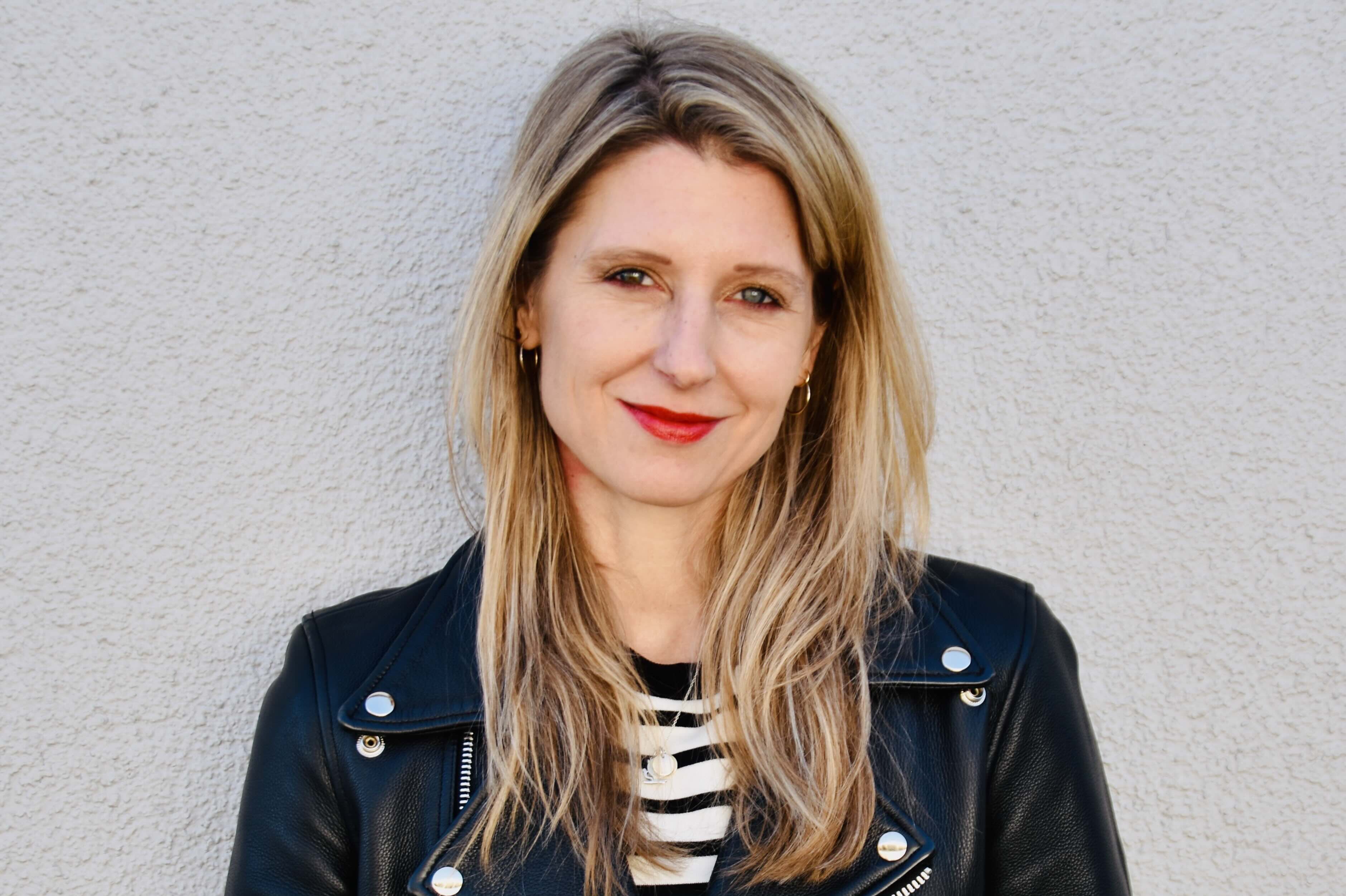 Laura Fröhlich, Redakteurin, Porträt mit warmem Lächeln und direktem Blick – schulterlange braune Haare mit Highlights, roter Lippenstift, schwarze Lederjacke über Streifenshirt, professionell und nahbar, Vertrauen und Expertise