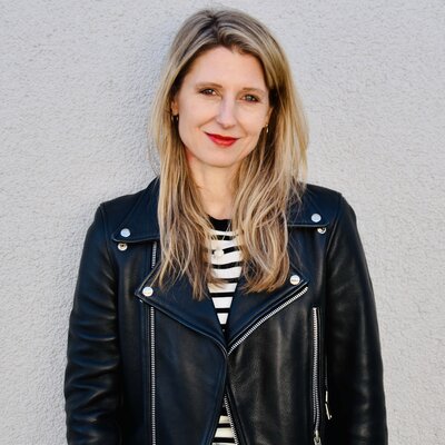 Laura Fröhlich, Redakteurin, Porträt mit warmem Lächeln und direktem Blick – schulterlange braune Haare mit Highlights, roter Lippenstift, schwarze Lederjacke über Streifenshirt, professionell und nahbar, Vertrauen und Expertise