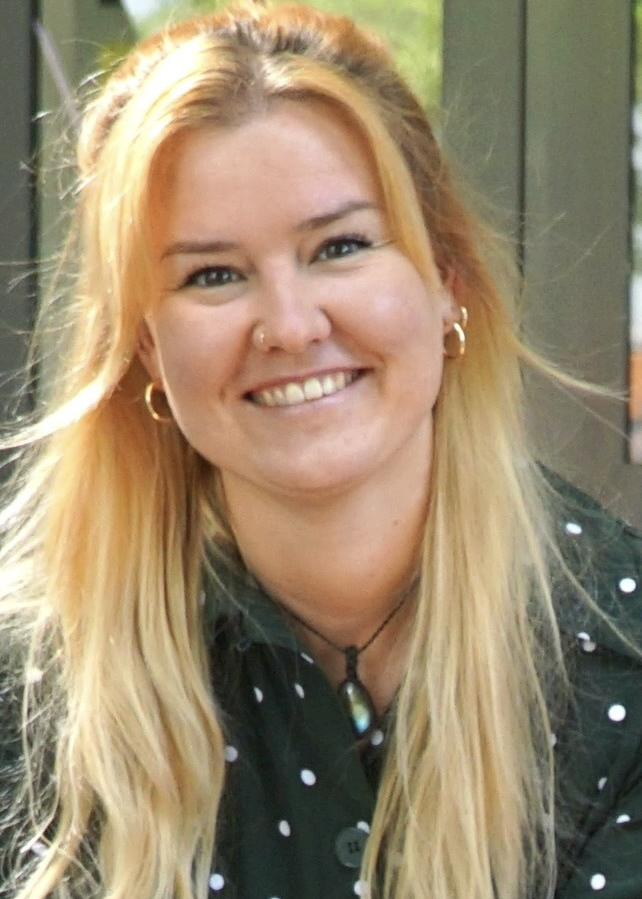 Melanie Lachmann, Redakteurin, Porträt mit warmem Lächeln und direktem Blick – lange rotgoldblonde Haare mit Wellen bis zur Schultermitte, blaugraue Augen, dunkelgrüne Bluse mit weißem Polka-Dot-Muster, goldene Ohrringe, sitzt entspannt mit gefalteten Händen, vertrauenswürdig und nahbar, moderne Außenkulisse mit grüner Vegetation im Hintergrund