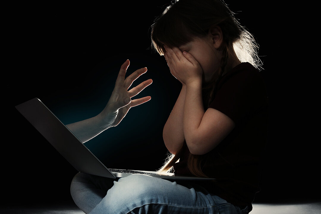 Kind vor Laptop, verdeckt das Gesicht, symbolisch für Online-Gefahren.