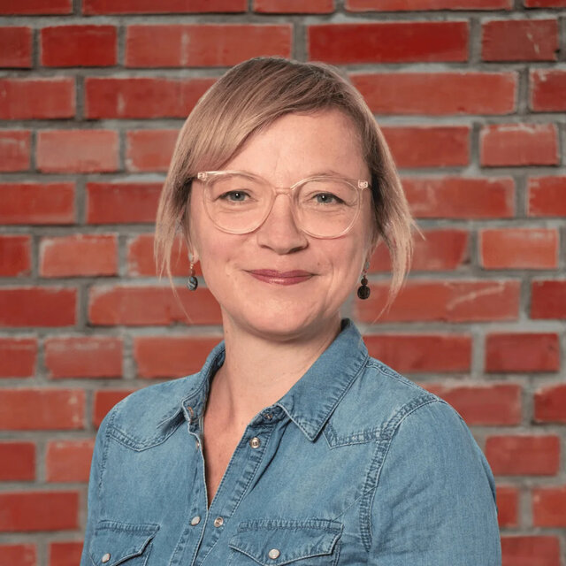 Porträt von Meike Wiggers, Gastautorin bei BARMER Durchblickt, mit blonden Haaren und Brille vor roter Backsteinwand.