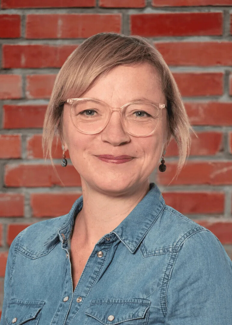 Porträt von Meike Wiggers, Gastautorin bei BARMER Durchblickt, mit blonden Haaren und Brille vor roter Backsteinwand.