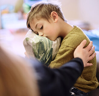 Ein Junge sitzt traurig auf einem Sofa und hält ein Kissen im Arm. Eine erwachsene Person legt ihm tröstend die Hand auf die Schulter.