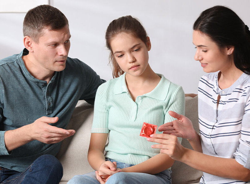 Zwei Erwachsene sprechen mit einem Mädchen auf dem Sofa und erklären ihr anhand eines Kondoms das Thema Verhütung
