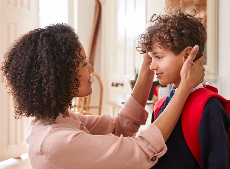 Mutter richtet ihrem Sohn mit Schulrucksack im Eingangsbereich liebevoll die Haare, bevor er das Haus verlässt.