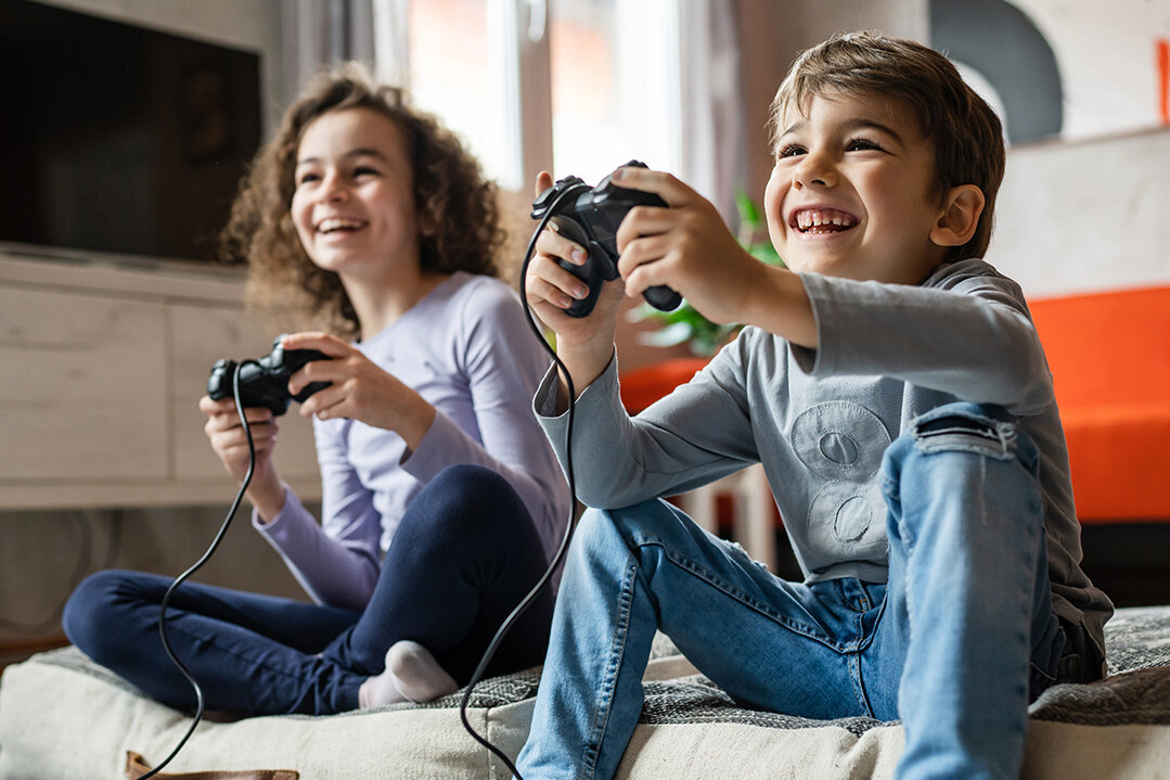 Junge und Mädchen spielen lachend gemeinsam Videospiele mit Controllern im hellen Wohnzimmer.
