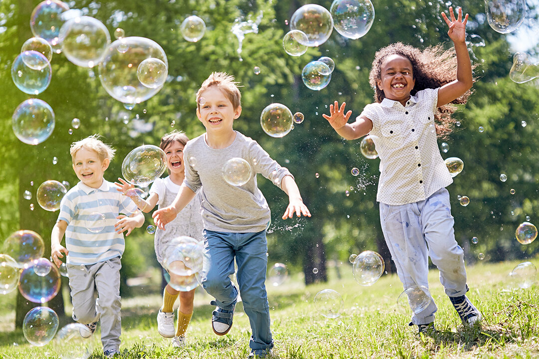 Vier Kinder rennen lachend über eine Wiese und spielen mit Seifenblasen.
