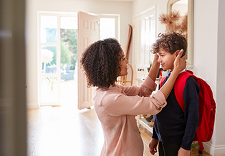 Mutter richtet ihrem Sohn mit Schulrucksack im Eingangsbereich liebevoll die Haare, bevor er das Haus verlässt.