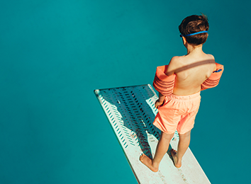 Junge mit Schwimmflügeln steht auf Sprungbrett im Schwimmabd und schaut ins Beclken.