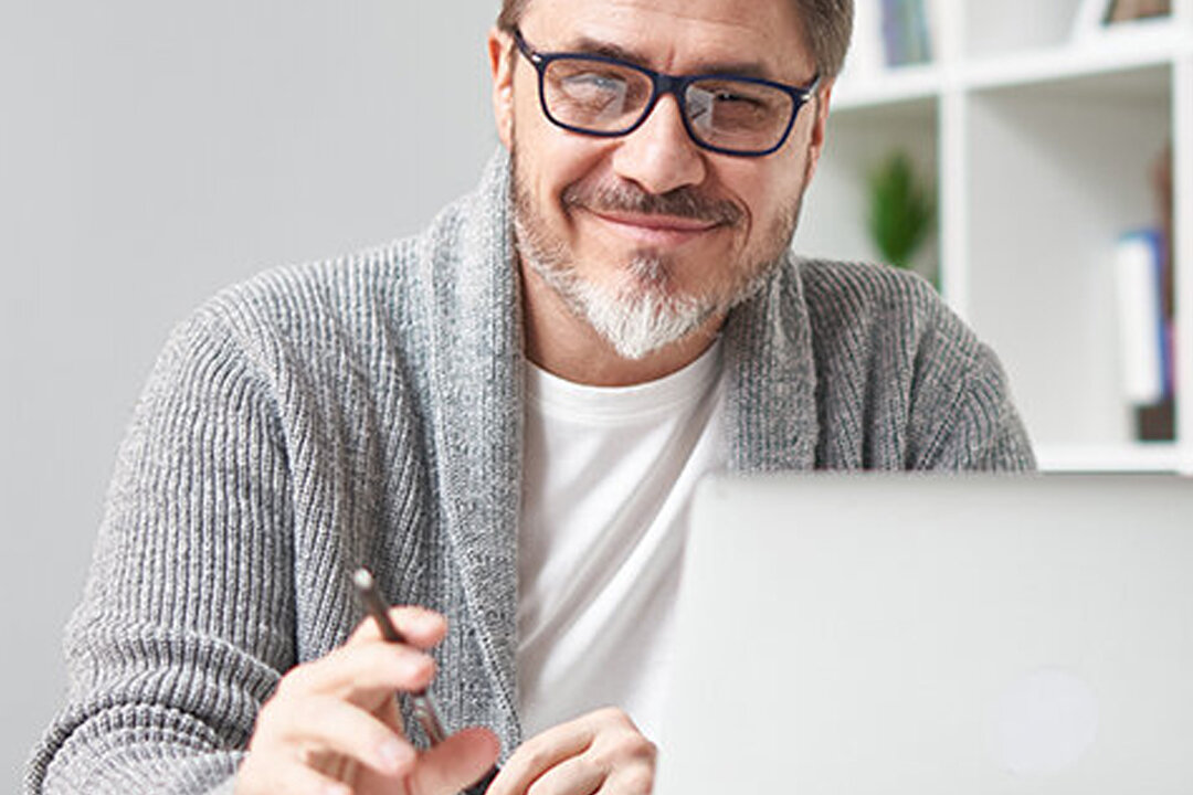 Mann mit Brille und grauer Strickjacke sitzt vor Laptop und blickt lächelnd in die Kamera