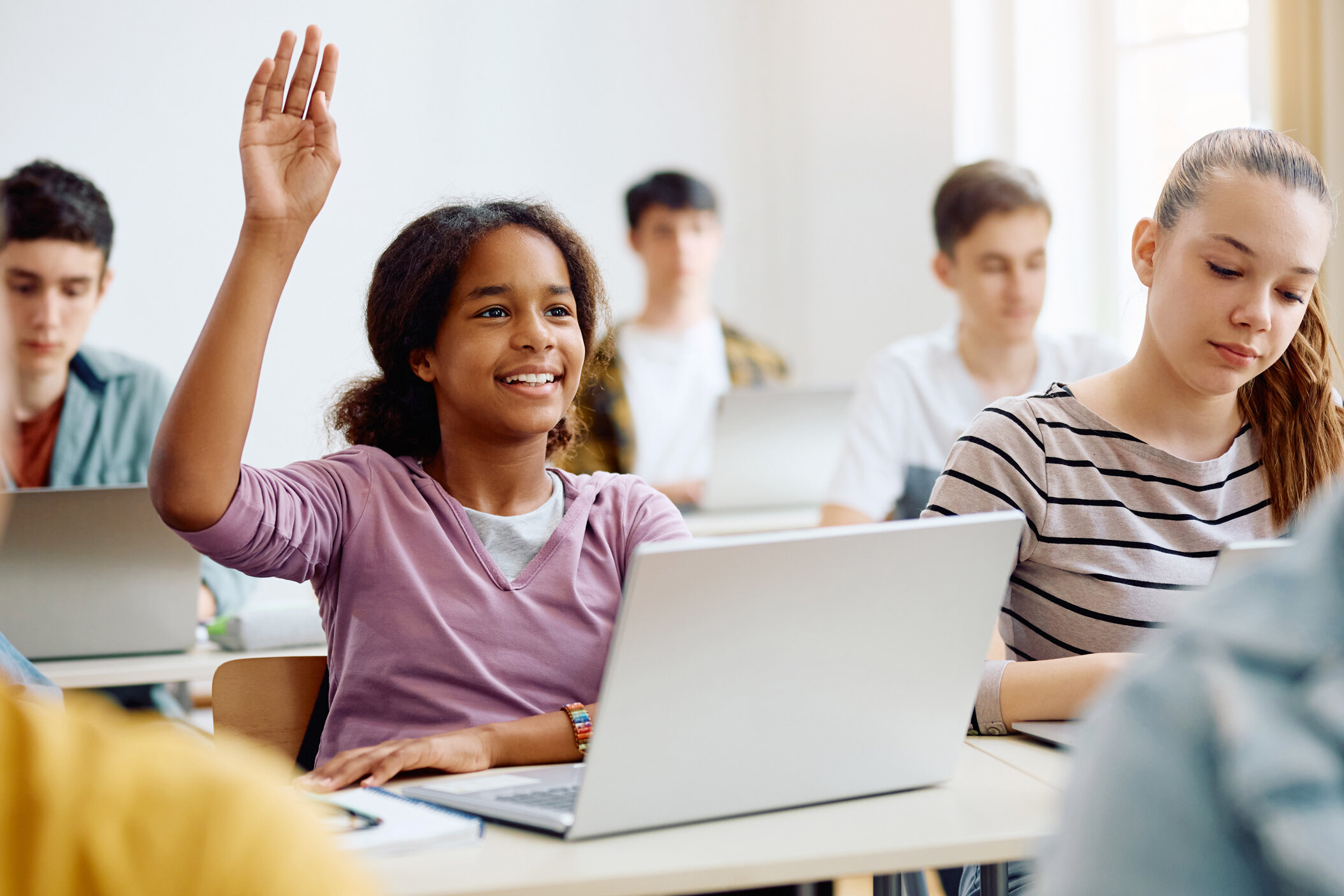 Schülerin mit Regenbogenarmband hebt Hand am Laptop-Schreibtisch, lächelt engagiert