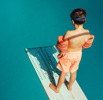 Junge mit Schwimmflügeln steht auf Sprungbrett im Schwimmabd und schaut ins Beclken.
