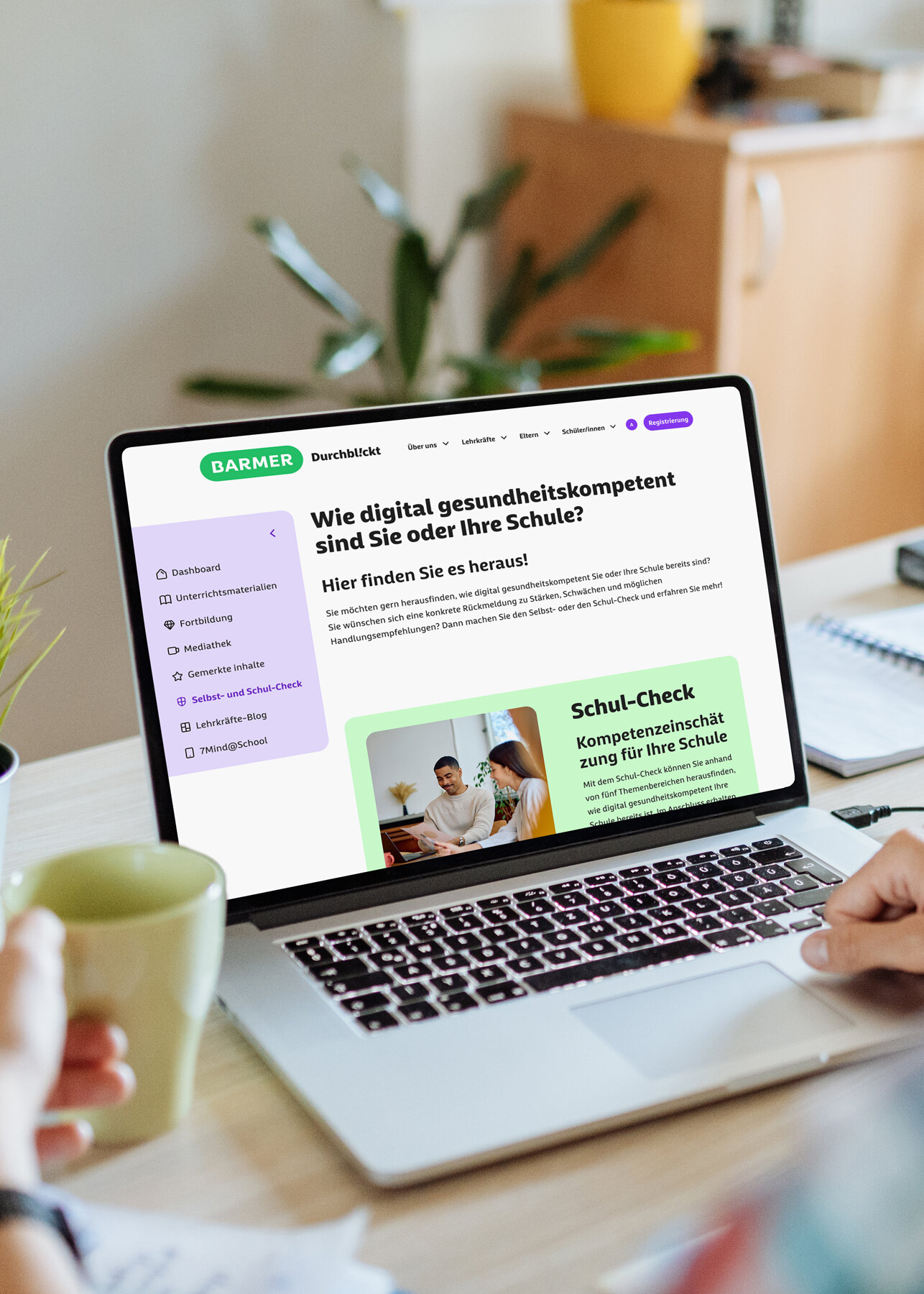 Lehrerin am Holzschreibtisch mit Laptop zeigt BARMER Durchblickt Schul-Check zur digitalen Gesundheitskompetenz, grüne Buttons, moderne Schulumgebung