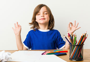 Kind sitzt mit geschlossenen Augen am Tisch und meditiert, daneben liegen bunte Stifte und Papier