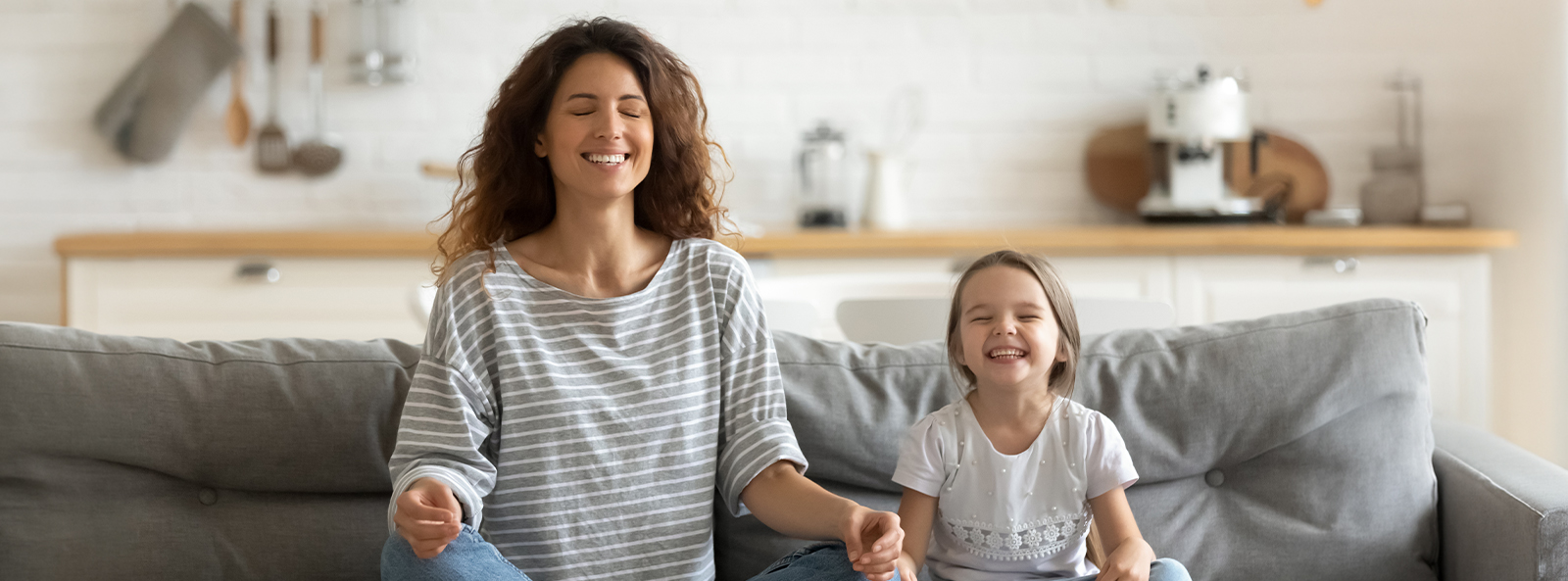 Mutter und Tochter praktizieren gemeinsam Achtsamkeit und Meditation zu Hause – beide sitzen entspannt im Schneidersitz auf dem Sofa mit geschlossenen Augen