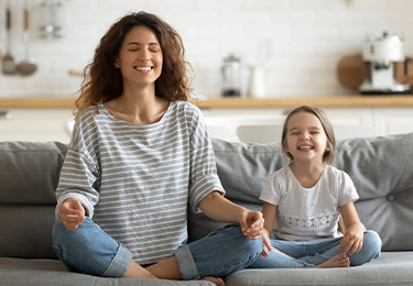 Mutter und Tochter praktizieren gemeinsam Achtsamkeit und Meditation zu Hause – beide sitzen entspannt im Schneidersitz auf dem Sofa mit geschlossenen Augen