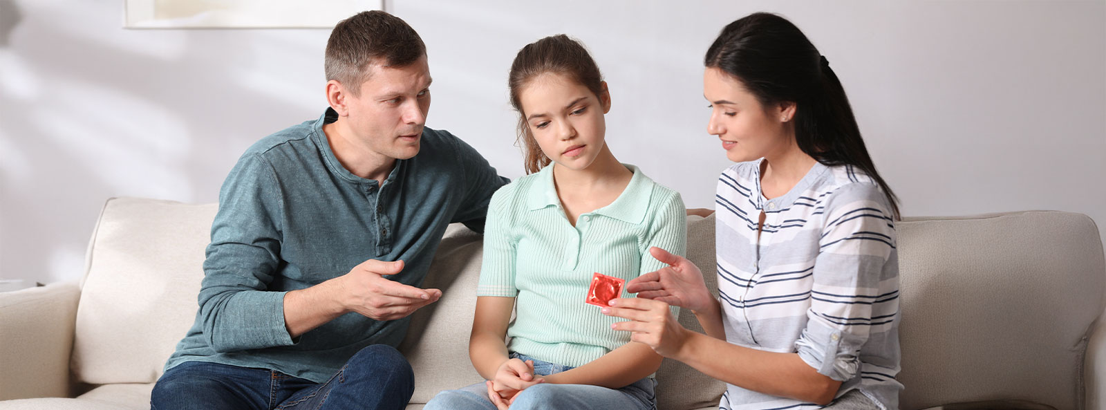 Zwei Erwachsene sprechen mit einem Mädchen auf dem Sofa und erklären ihr anhand eines Kondoms das Thema Verhütung