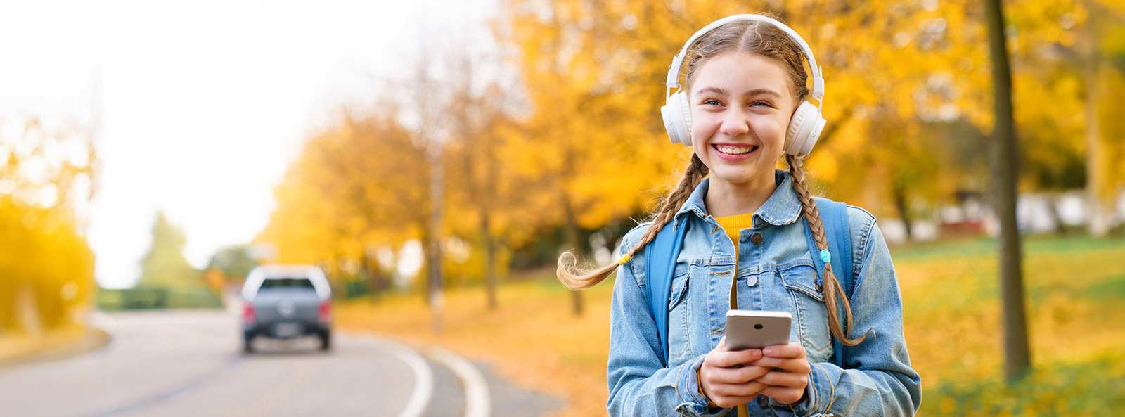 Ein lächelndes Mädchen mit Kopfhörern geht im Herbst an einer Strasse entlang. Sie hat ein Smartphone in der Hand.
