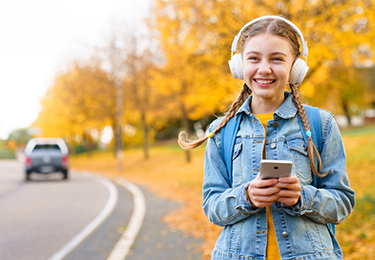 Ein lächelndes Mädchen mit Kopfhörern geht im Herbst an einer Strasse entlang. Sie hat ein Smartphone in der Hand.