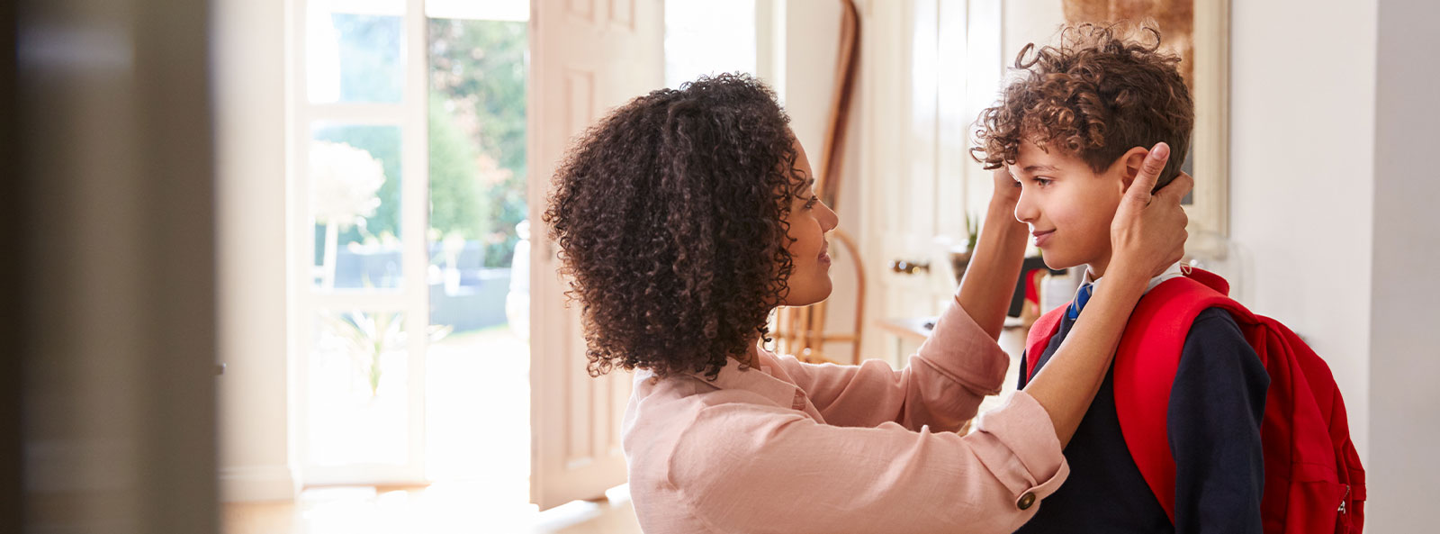 Mutter richtet ihrem Sohn mit Schulrucksack im Eingangsbereich liebevoll die Haare, bevor er das Haus verlässt.