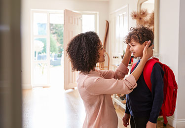 Mutter richtet ihrem Sohn mit Schulrucksack im Eingangsbereich liebevoll die Haare, bevor er das Haus verlässt.