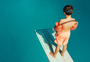 Junge mit Schwimmflügeln steht auf Sprungbrett im Schwimmabd und schaut ins Beclken.
