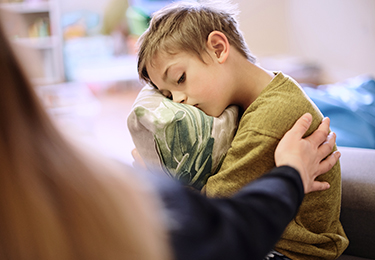 Ein Junge sitzt traurig auf einem Sofa und hält ein Kissen im Arm. Eine erwachsene Person legt ihm tröstend die Hand auf die Schulter.