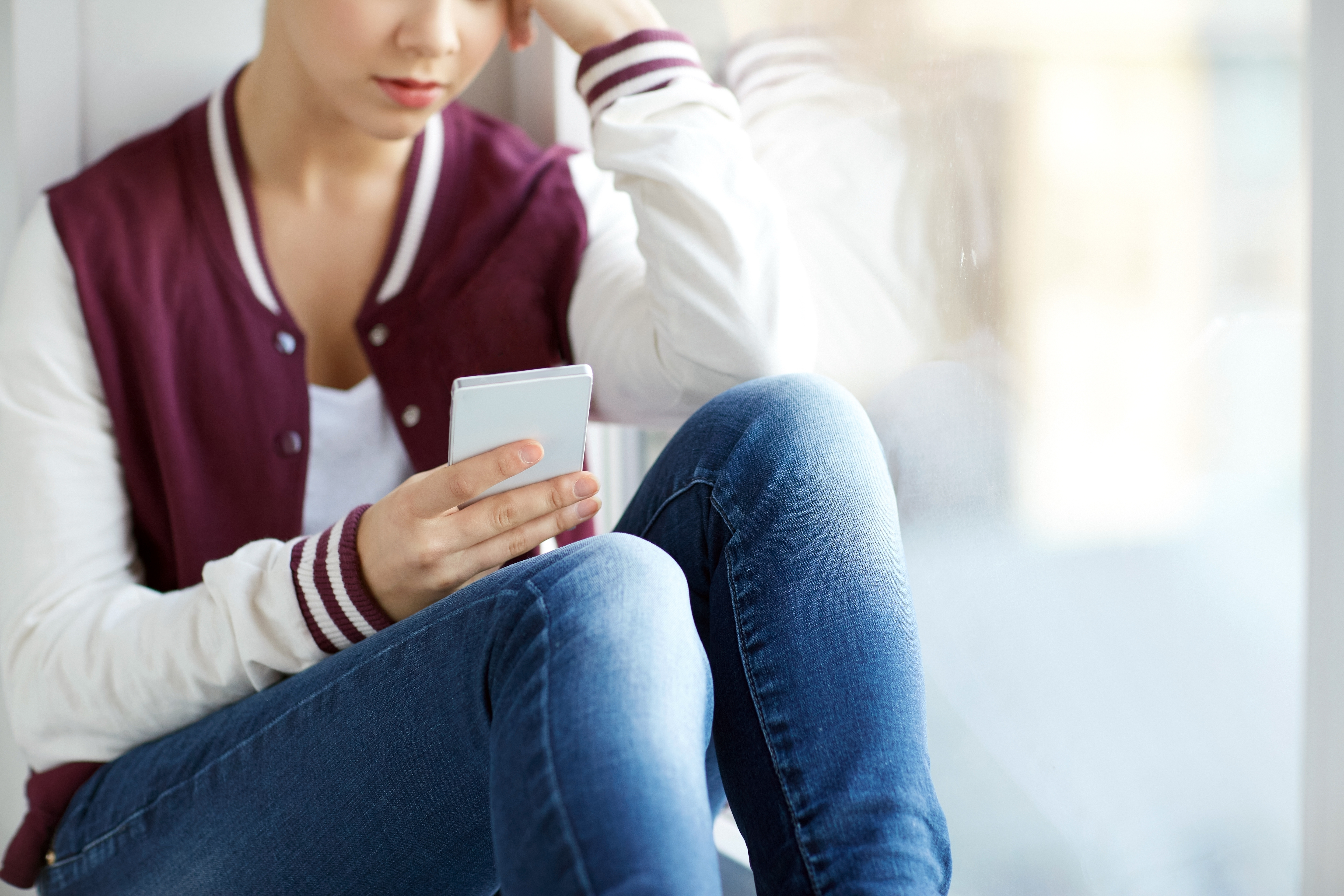 Person sitzt mit angezogenen Beinen auf dem Boden und sieht nachdenklich auf Smartphone