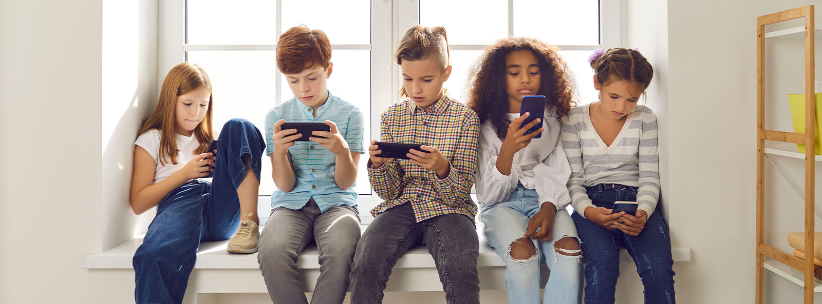 Fünf Kinder sitzen nebeneinander auf einer Fensterbank und schauen konzentriert auf ihre Smartphones.