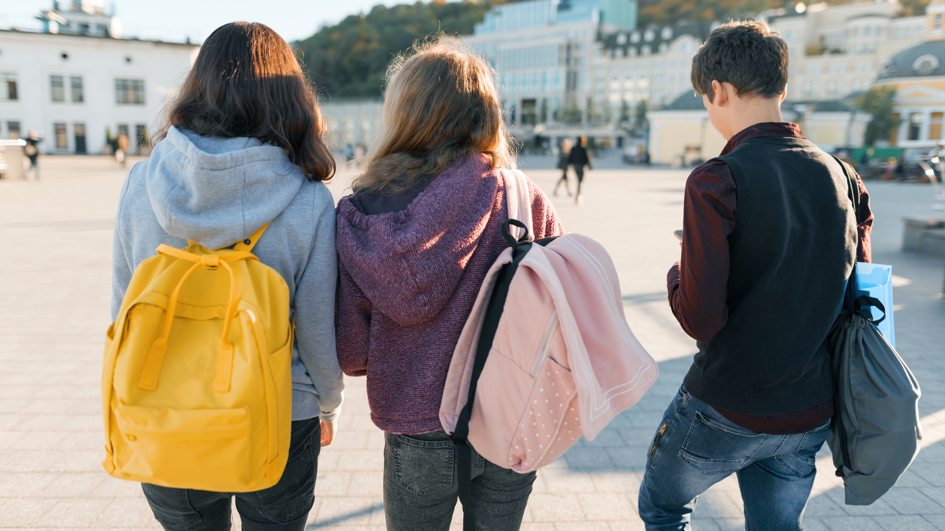 3 Teenager laufen über einen Platz. Man sieht sie von hintern. Sie tragen Rucksäcke.