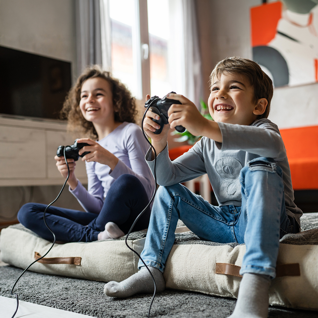 Junge und Mädchen spielen lachend gemeinsam Videospiele mit Controllern im hellen Wohnzimmer. BARMER-Durchblickt-artikel-eltern-dark-patterns.jpg