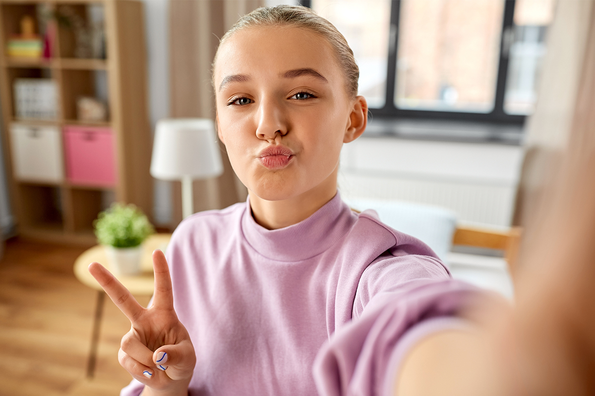 Eine junge Frau macht lächelnd ein Selfie in einem hellen Wohnzimmer. Sie trägt einen rosafarbenen Pullover, zeigt ein Peace-Zeichen und blickt entspannt in die Kamera. Das Bild illustriert den selbstbewussten und spielerischen Umgang mit sozialen Medien und Selbstdarstellung.