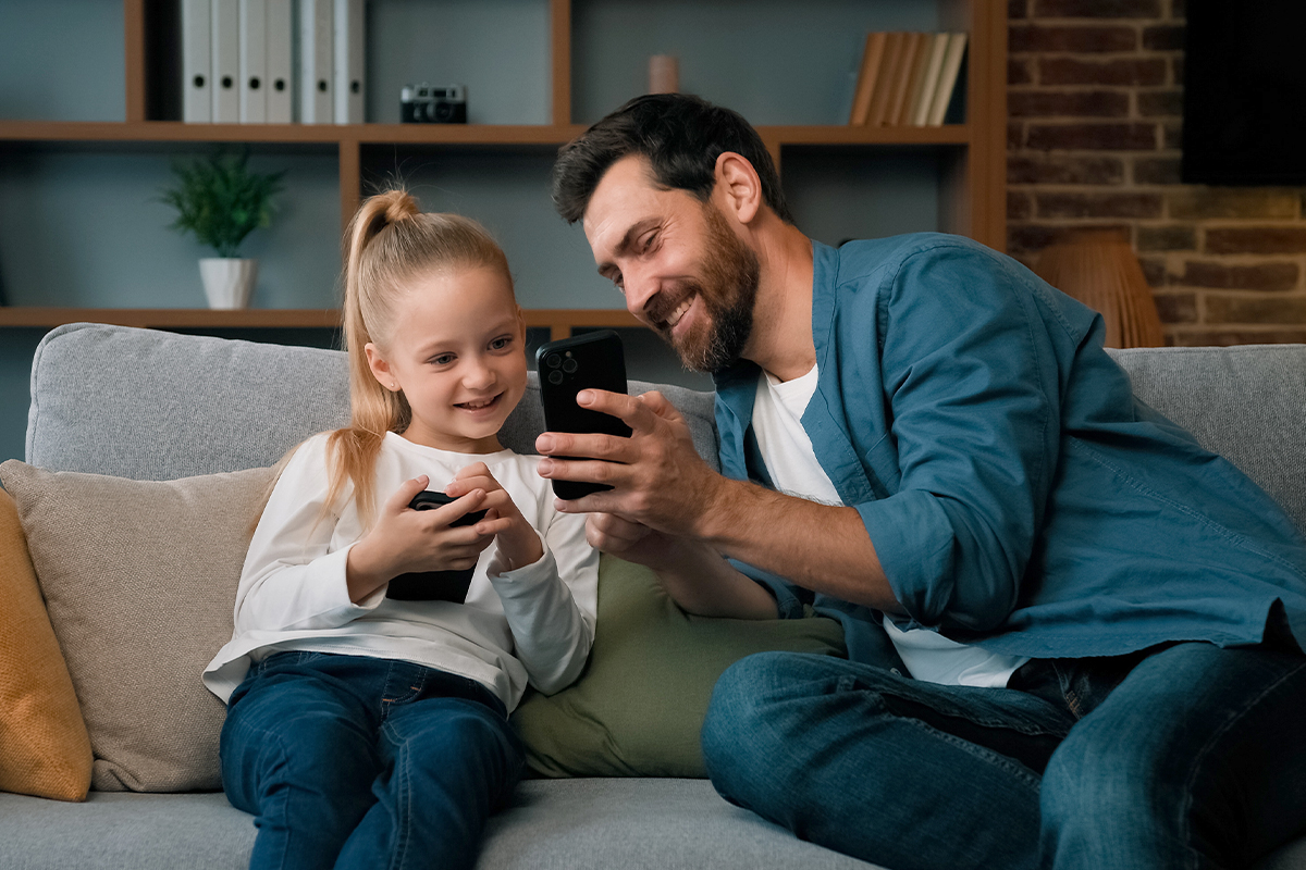  Ein Mann zeigt einem Mädchen etwas auf seinem Smartphone. Beide halten ein Gerät in der Hand und sitzen auf einem Sofa.