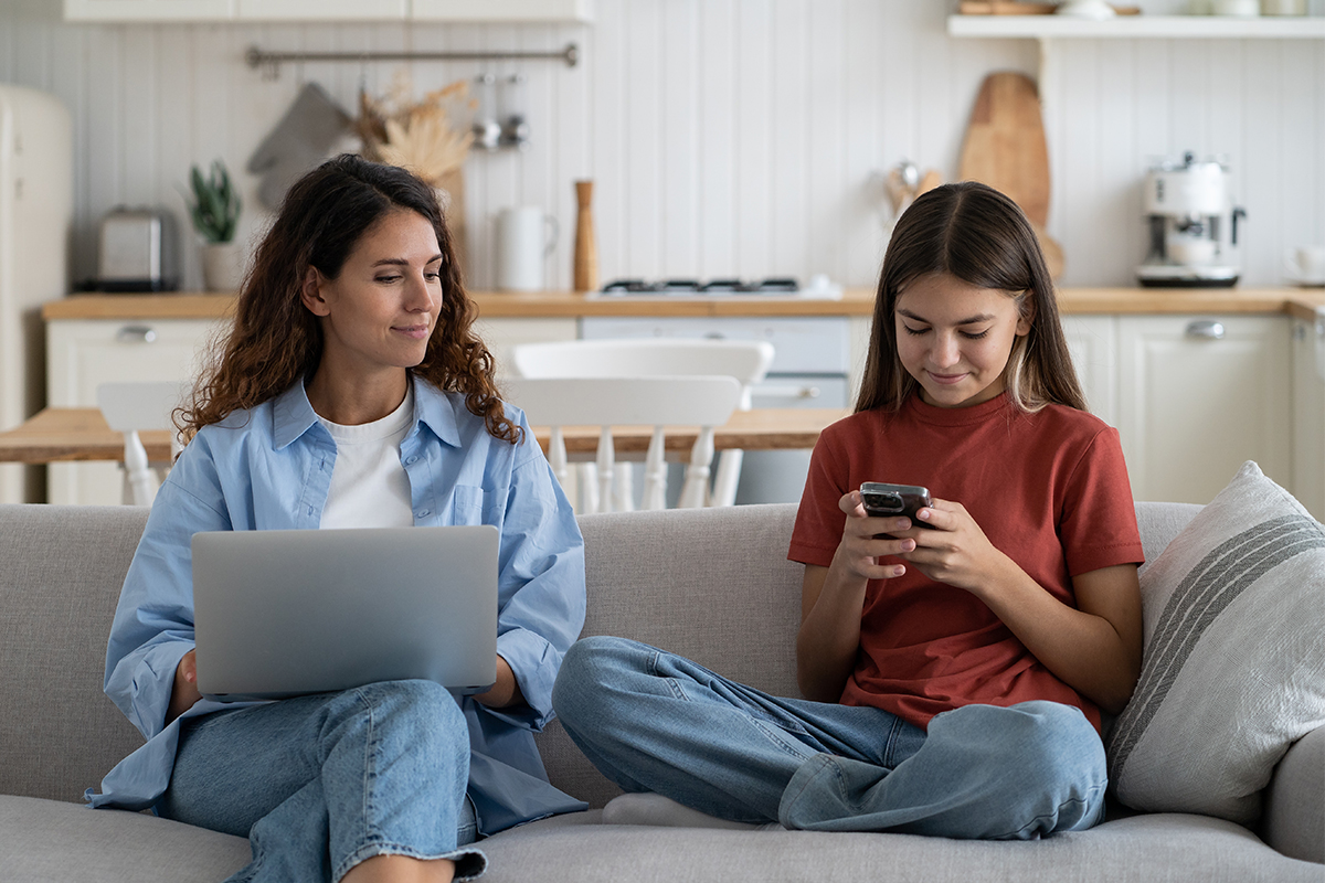 Eine Mutter am Laptop schaut ihrer Tochter zu, wie sie am Smartphone ist. Sie Sitzen auf dem Sofa.
