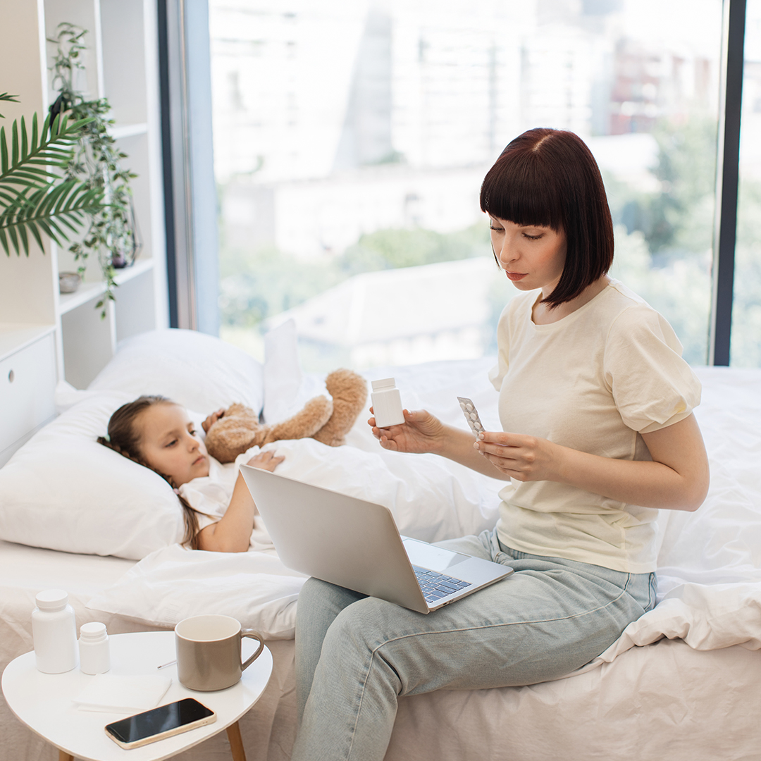 Kind liegt krank im Bett. Mutter sitzt daneben, hat Tablettenblister in der Hand un dschaut auf einen Laptop 20260526_DGK_Eltern_Artikel_Dr-Chat-GPT.jpg
