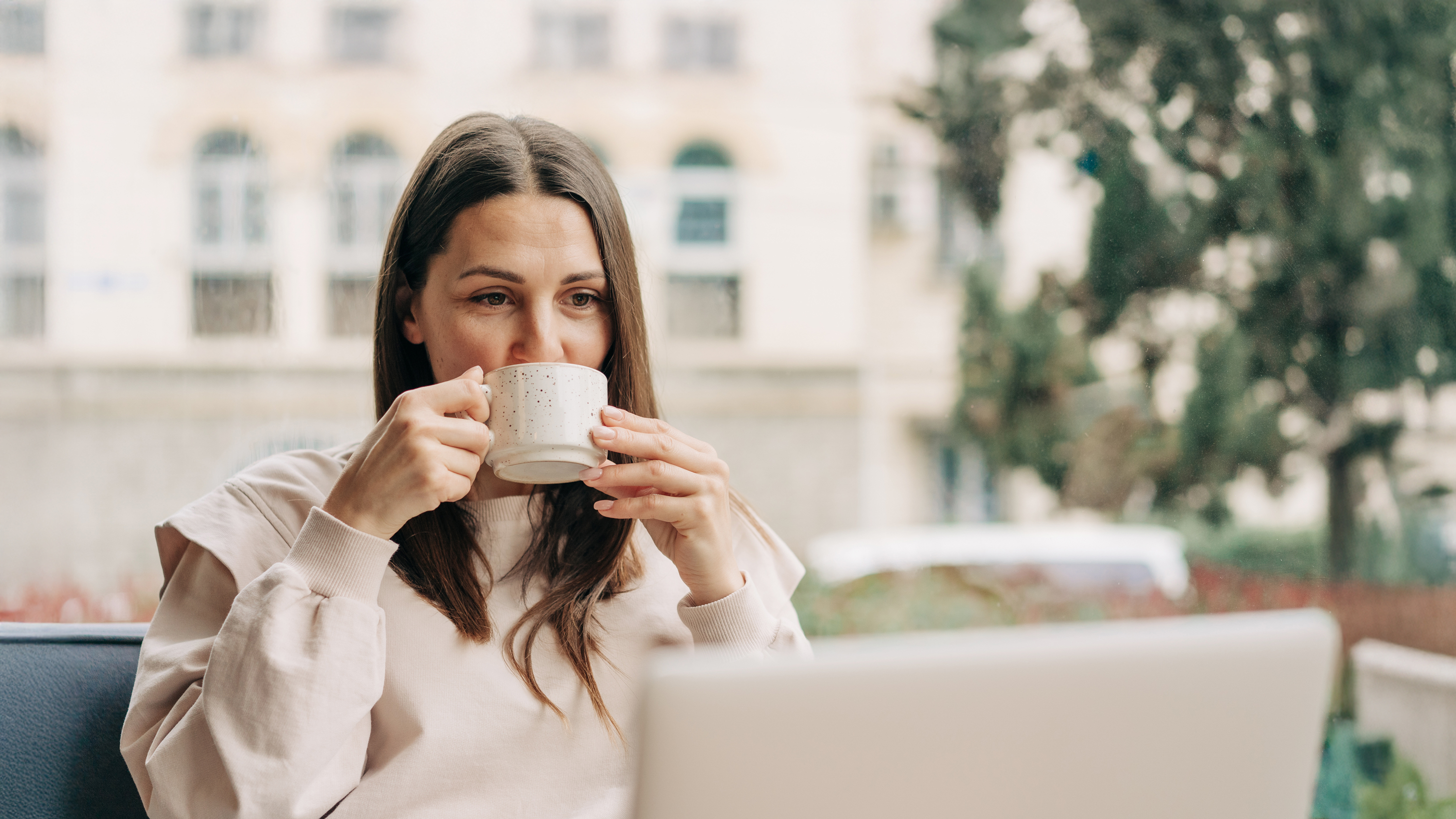 Junge Frau mit langen braunen Haaren sitzt an einem Laptop und trinkt aus einer beigen Keramiktasse mit Punktmuster, während sie entspannt in die Kamera schaut – Homeoffice, Work-Life-Balance, digitale Gesundheit, Produktivität