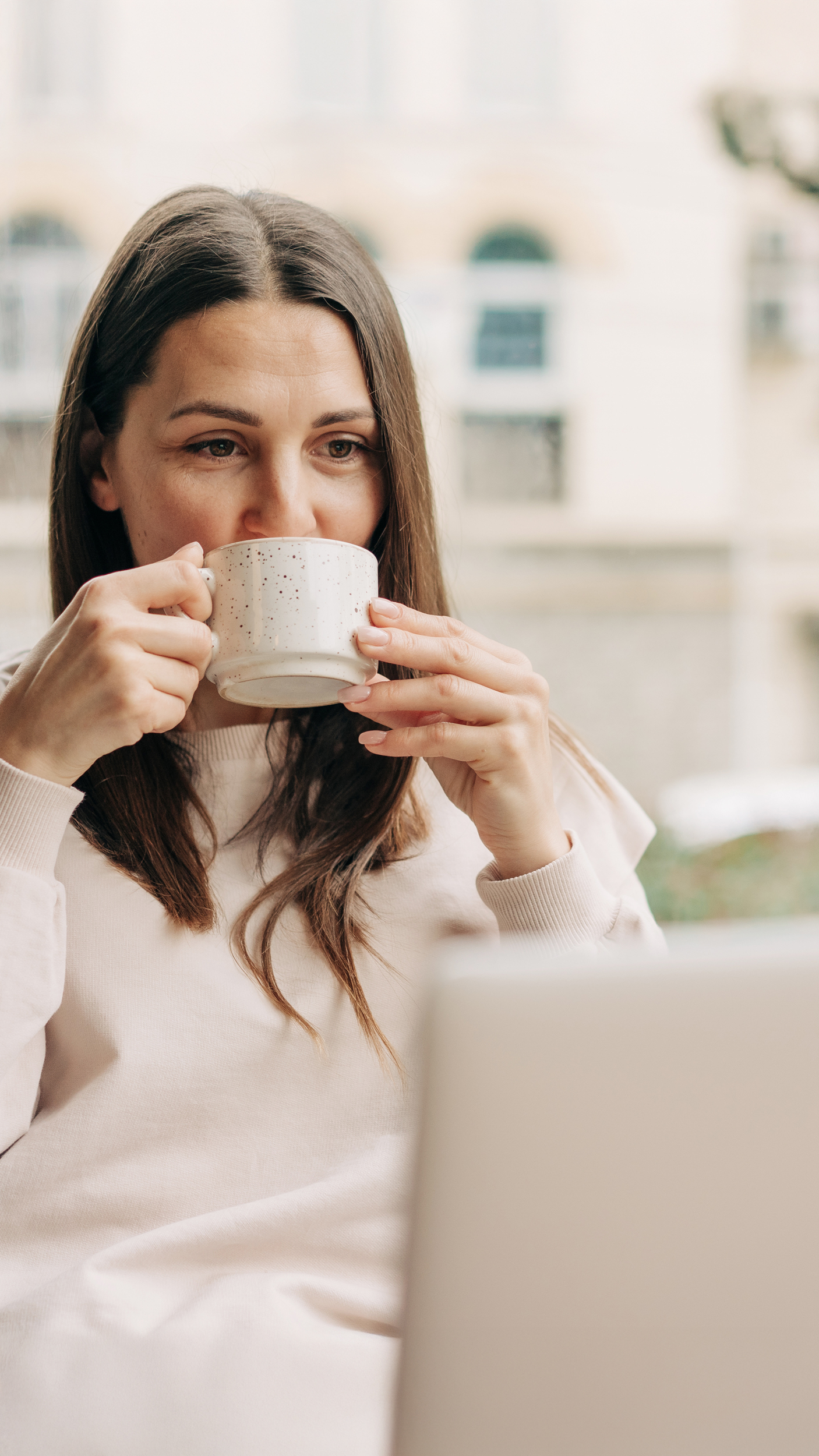 Junge Frau mit braunen Haaren sitzt an einem Laptop und trinkt aus einer Tasse während sie entspannt blickt – Homeoffice, Work-Life-Balance, digitale Gesundheit, Produktivität